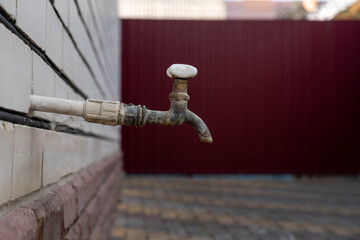 Outdoor plumbing tap with weathered metal faucet stands on tiled wall, framed in close up against blurred red background. Outdoor plumbing tap shows aged surfaces and no water flow. © Egor Lint