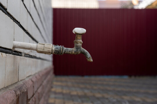 Outdoor plumbing tap with weathered metal faucet stands on tiled wall, framed in close up against blurred red background. Outdoor plumbing tap shows aged surfaces and no water flow.