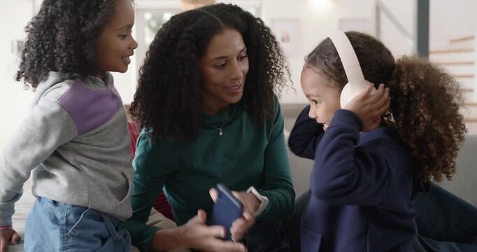 African American mom with daughters taking white headphones and checking phone for music on couch