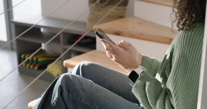 African American adult woman in green knit tapping phone prompted by phone on stairs, reading texts