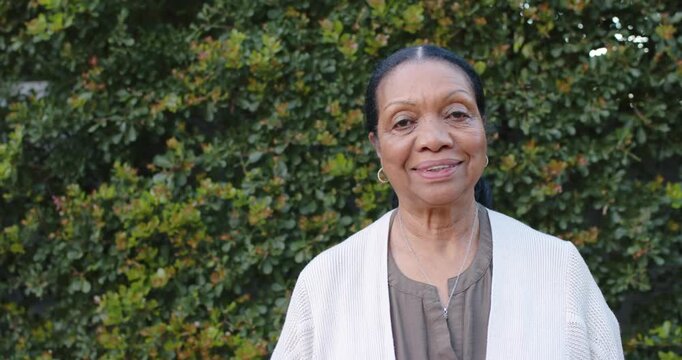 Senior African American woman noticing camera, smiling, laughing, leaning at hedge wearing cardigan