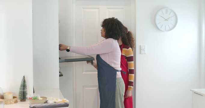 African American mother guiding youth daughter sliding oven rack after timer in kitchen in aprons
