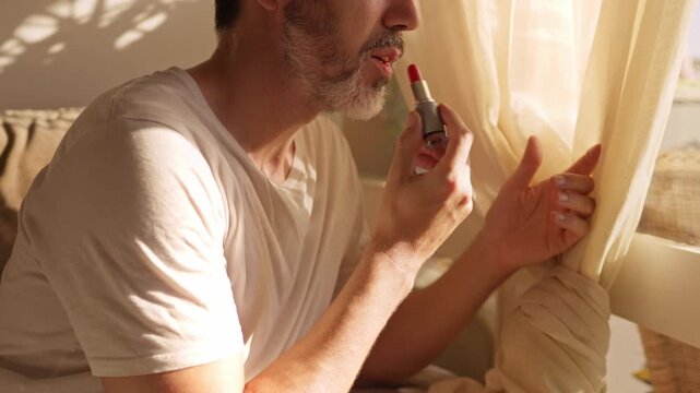 queer person with a beard applying makeup by a window with warm sunlight