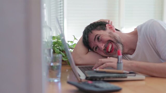 Genderfluid person with beard and red lipstick laughing while working from home on a laptop