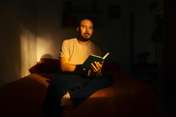 Relaxed man reading a book while sitting cross-legged on a bed, smiling contentedly in warm sunlight. Dramatic shadows and a cozy bedroom create a calm, intimate atmosphere. © Francis