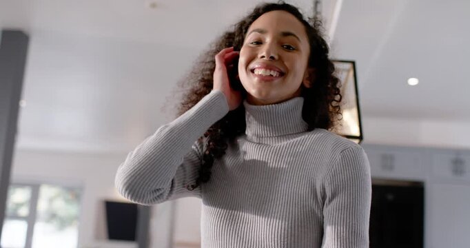 African American woman standing in open-plan home lifting phone, recording selfie and tucking hair