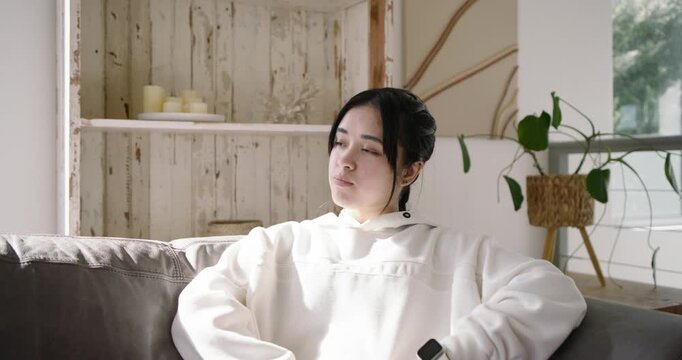 Asian female reacting to sunlight, shifting gaze toward camera, smiling laughing on gray couch
