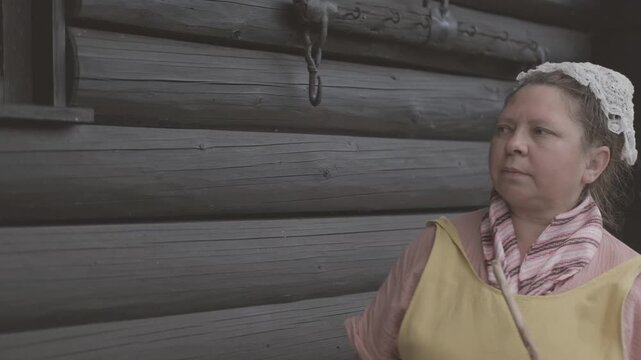 woman in traditional peasant costume, yellow pinafore dress, pink blouse, patterned scarf, lace headpiece, Historical portrait, holding wooden stick, near dark log cabin wall, basket, rural heritage