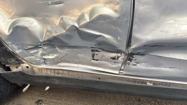 Close up of a severely dented silver car door with deep scratches, damaged metal and visible impact marks