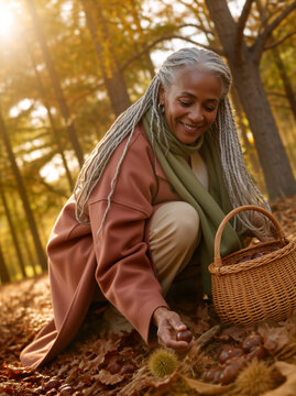 Senior black woman gathering chestnuts in an autumn forest. Older female with grey braids picking up nuts from fallen leaves into a wicker basket