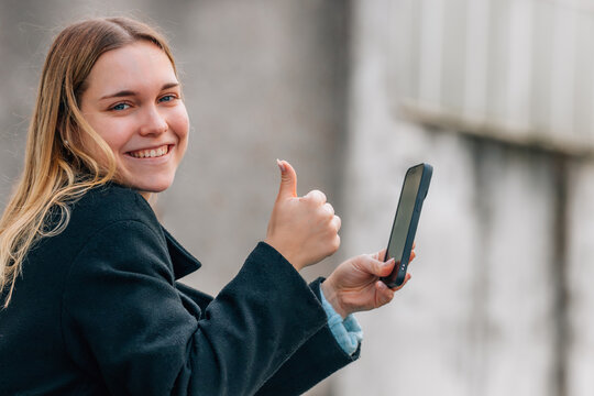 Happy young Caucasian woman giving a thumbs up while holding a smartphone outdoors, smiling with a positive and cheerful expression