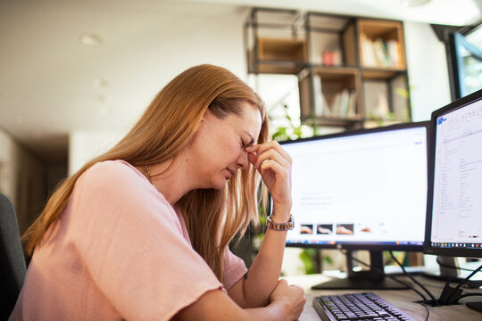 Stressed woman rubbing eyes at office desk with computer monitors