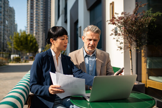 Two business colleagues reviewing documents on laptop at outdoor cafe