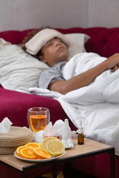 Teenage boy resting in bed with a cold compress on his forehead, surrounded by tissues, citrus fruit, warm tea, nasal sprays, and medicine, representing seasonal illness, common cold symptoms, fever