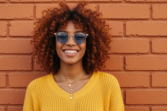 Smiling trans woman with curly afro hair wears trendy sunglasses, yellow sweater. Happy hispanic lady posing by red brick wall in city. Fashionable person shows pride, identity with confident look,