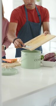Vertical video: Couple at kitchen island, man scraping board into green bin after cutting carrots