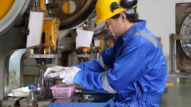 Industrial worker operating metal press machine in factory workshop, wearing protective helmet, glove, blue coverall during manufacturing production and precision metal production work