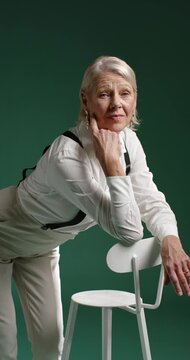 Vertical video: Senior woman in studio reacting on cue leaning on white chair in suspenders posing