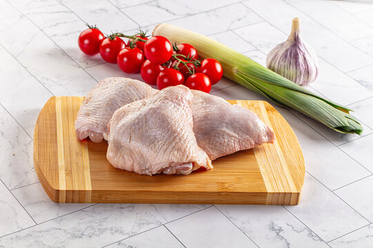 Raw chicken thighs on wooden cutting board with cherry tomatoes, leek, and garlic on marble countertop