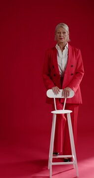 Vertical video: Mature woman in red suit holding stool in studio turning to camera after glance