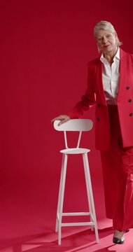 Vertical video: Senior woman in red suit touching stool stepping to center sitting posing in studio