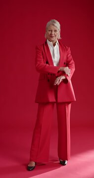 Vertical video: Senior woman posing on red studio floor, looking down then adjusting blazer
