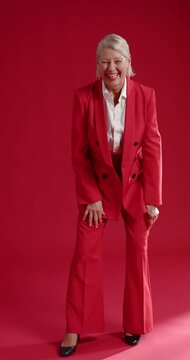 Vertical video: Senior female reacting to camera cue, laughing and gesturing in red suit in studio