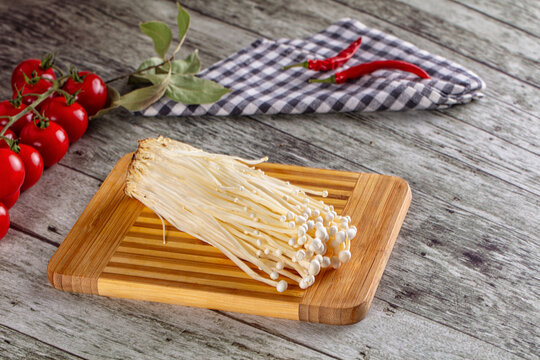 Fresh enoki mushrooms on wooden cutting board with cherry tomatoes, bay leaves, and red chili peppers on rustic wood background