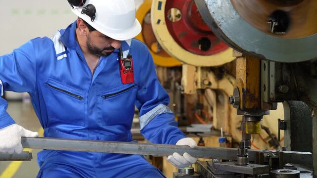 Industrial worker operating metalworking machine factory, blue uniform, white helmet, safety glasses, beard, male adult wearing protective gear while handling steel rod during metal manufacturing