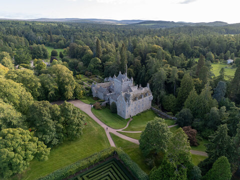 High wide static aerial photograph over Cawdor Castle with its stone tower, manicured lawns, and surrounding lush green forest near Nairn, Scotland