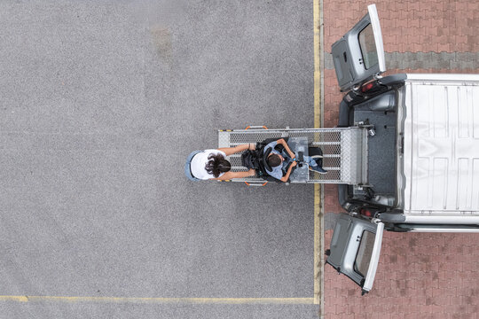 Personal assistant helping a wheelchair user enter an adapted vehicle using an accessible ramp, aerial view.