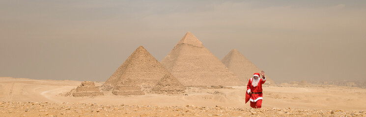 Naklejka premium Playful Santa Claus figure in red suit waving on the sandy Giza desert plateau with the iconic Great Pyramids towering in the background under a hazy