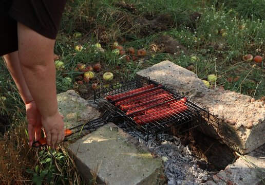 Person holding a wire grill basket with sausages over hot coals in a stone pit
