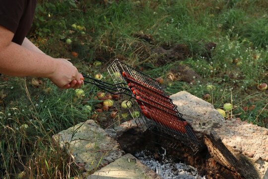 Person holding a wire grill basket with sausages over hot coals in a stone pit