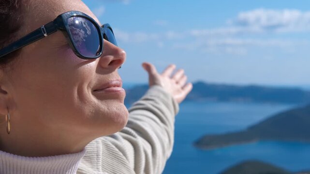 Happy woman feeling the sun on her face at a viewpoint. Close-up of a smiling woman in sunglasses with her arm outstretched, enjoying the warmth of the sun and a beautiful sea view, feeling free