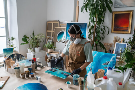 Woman wear respirator and protective gloves applies colored epoxy resin to abstract panel in home loft workshop, creating unique handmade artwork to monetize artistic hobby through small business.