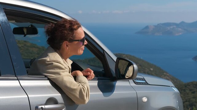 Woman traveler showing thumbs up from car window. Cheerful woman with sunglasses leaning out of a car window, admiring a stunning coastal view with mountains and the sea, then giving a thumbs up