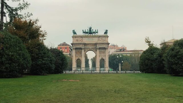 A scenic panning shot of the historic Arco della Pace triumphal arch with classical bronze statues and people relaxing in Piazza Sempione on a cloudy day in Milan.

