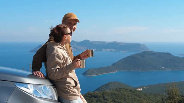 Couple enjoying coffee break during road trip with scenic sea view. Happy couple standing by their car, drinking hot tea from mugs while enjoying the breathtaking mountain and sea during road trip