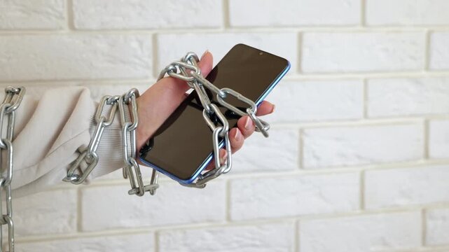 Digital slavery. Woman's hand wrapped in a heavy metal chain holding a smartphone against a white brick wall, symbolizing the concept of social media addiction, digital slavery, and nomophobia