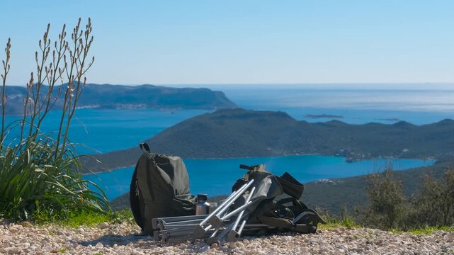 Hiker backpack and camping gear overlooking a beautiful sea bay. Camping equipment, including a backpack and a folding chair, resting on a gravel path on top with a stunning view of the turquoise sea