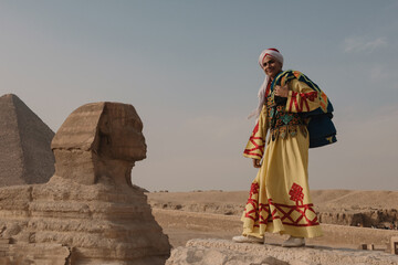 Naklejka premium Joyful man wearing ornate embroidered yellow robe and white turban stands smiling beside the Great Sphinx of Giza on the desert plateau ideal for