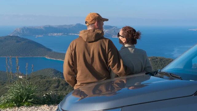 Romantic couple enjoying the sea view on a car road trip. Young loving couple standing together next to their car on a mountain road, hugging and enjoying the amazing scenic view of the blue sea