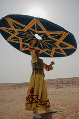 Naklejka premium Egyptian tanoura dancer in yellow embroidered robe raises enormous teal geometric spinning disc overhead in open sandy desert under blazing midday sun