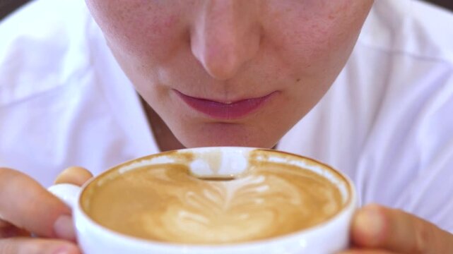 Young adult woman tastes latte coffee with concentrated expression, holding cup close to lips in cafe style closeup. Woman studies coffee aroma and foam while testing flavor with both hands.