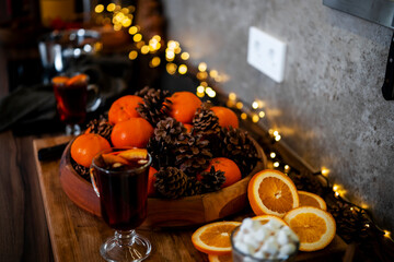 Naklejka premium Christmas hot wine drink and fresh oranges in wooden bowl — new year holiday preparation. Cozy winter evening kitchen interior featuring fairy lights. Festive seasonal decoration elements.