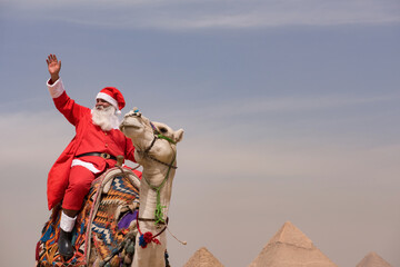 Naklejka premium Joyful Santa Claus in full red costume waving triumphantly while riding an ornately decorated camel near the Great Pyramids of Giza under open sky
