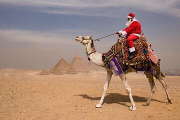 Naklejka premium Santa Claus in red costume rides a colorfully saddled camel mid stride across the Giza desert with iconic ancient pyramids behind under an overcast