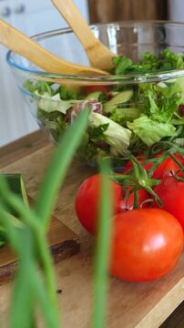 Mixed salad fills clear bowl as wooden spoons lean inside near chopped greens. Red tomatoes and tall green onions sit on wooden board, creating fresh farm style ingredients ready for serving.