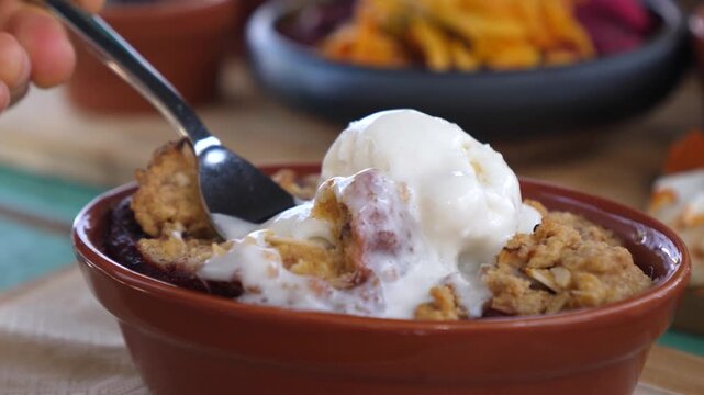 Vanilla ice cream and dessert crumble are stirred by spoon in close up, comforting sweet treat with rustic style and soft melted topping, served in brown ramekin over baked fruit, with syrup.